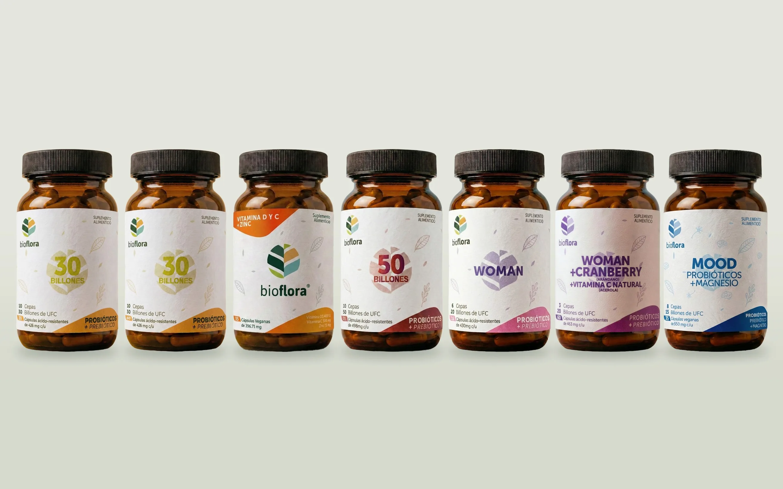 Los siete frascos de Bioflora en fila sobre un fondo gris verdoso claro: dos de 30 Billones, Vitamina D y C + Zinc, 50 Billones, Woman, Woman + Cranberry y MOOD, cada etiqueta en su color.