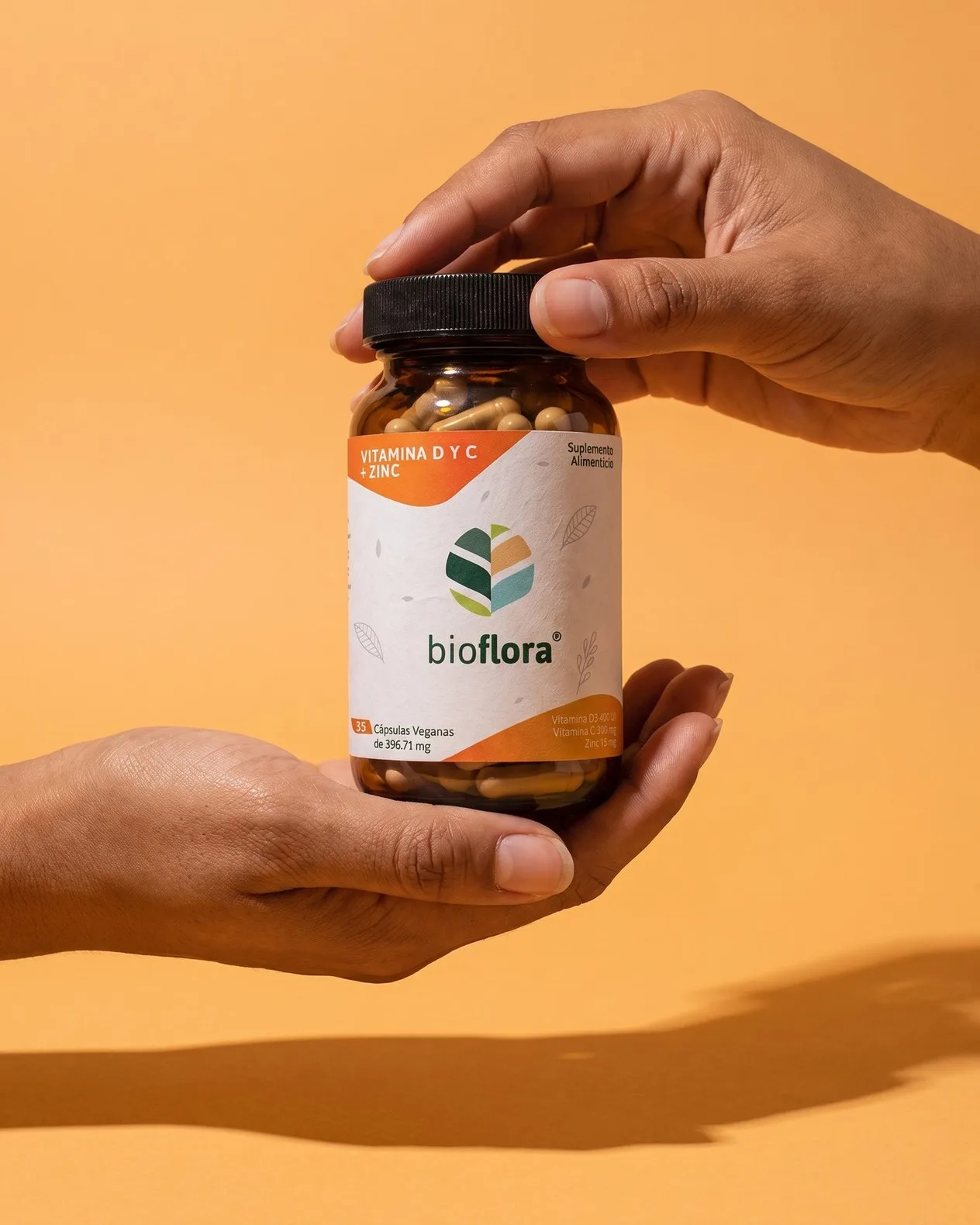 Dos manos sostienen un frasco de Bioflora Vitamina D y C + Zinc sobre un fondo naranja.