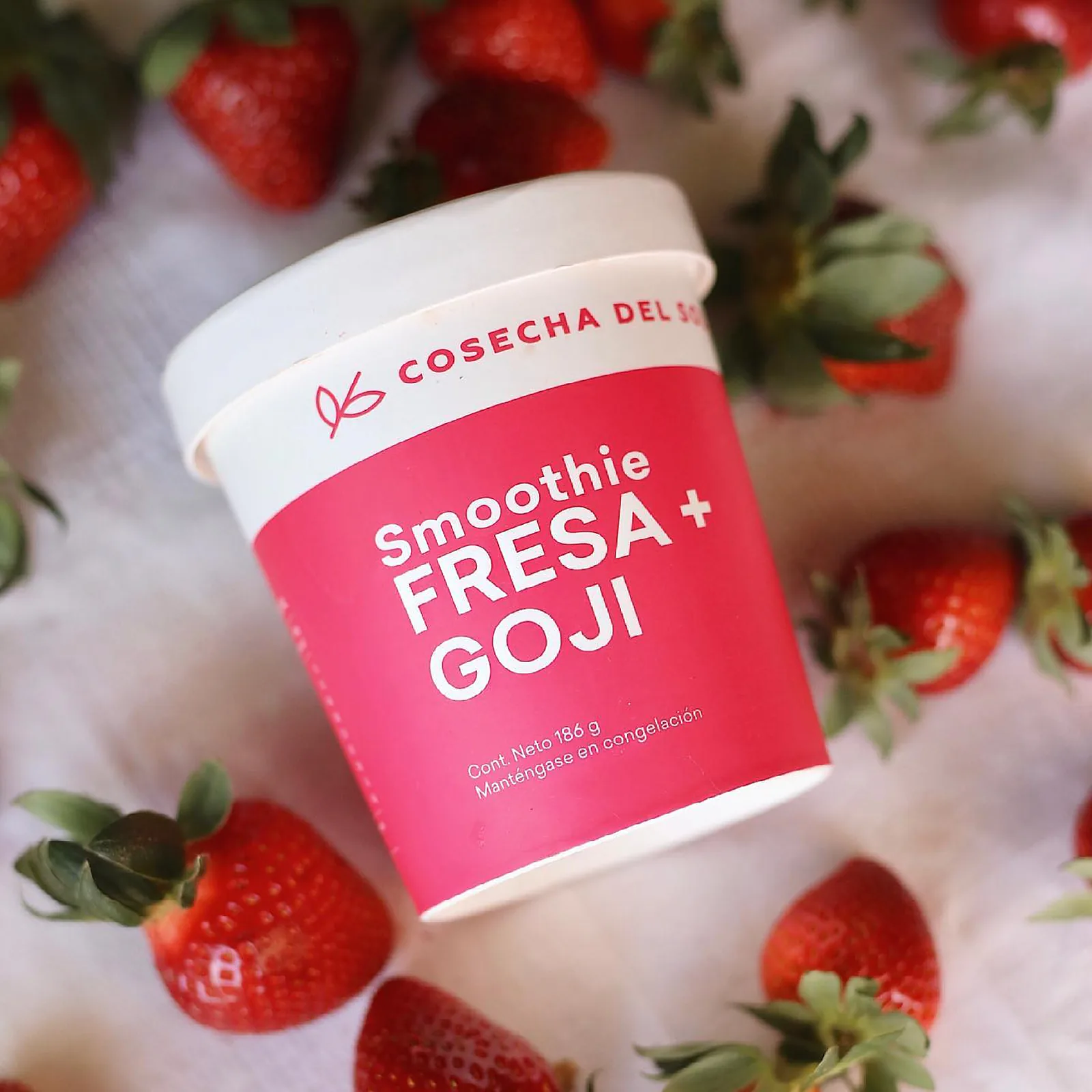Empaque rosa de smoothie Fresa + Goji entre fresas, sobre una tela blanca.