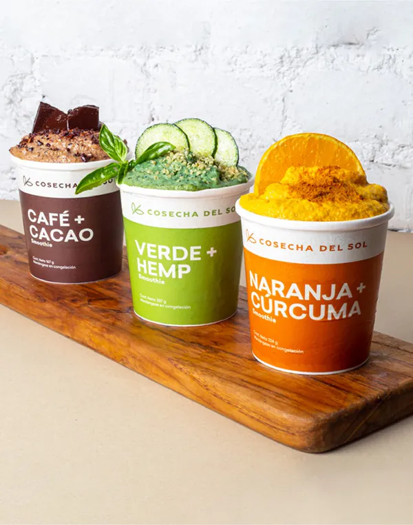 Tres empaques de smoothie, Café + Cacao, Verde + Hemp y Naranja + Cúrcuma, sobre una tabla de madera.