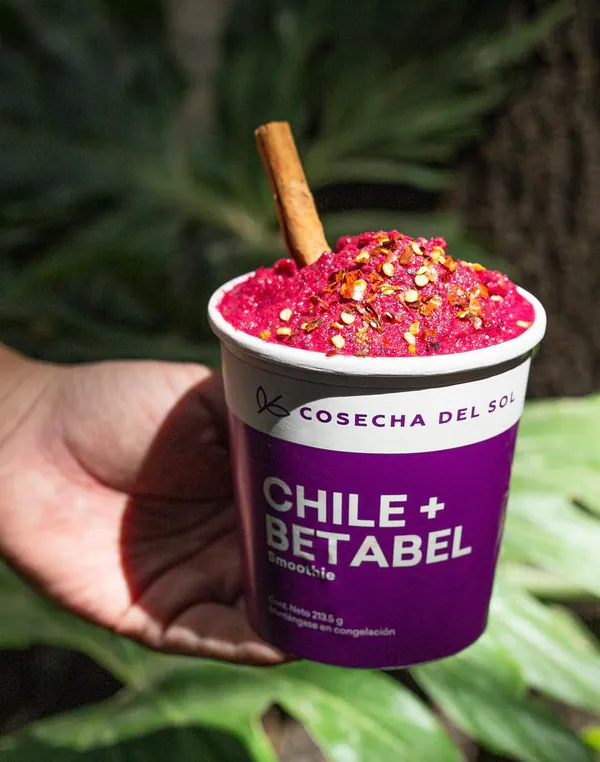 Mano que sostiene un empaque morado de Chile + Betabel frente a hojas verdes.