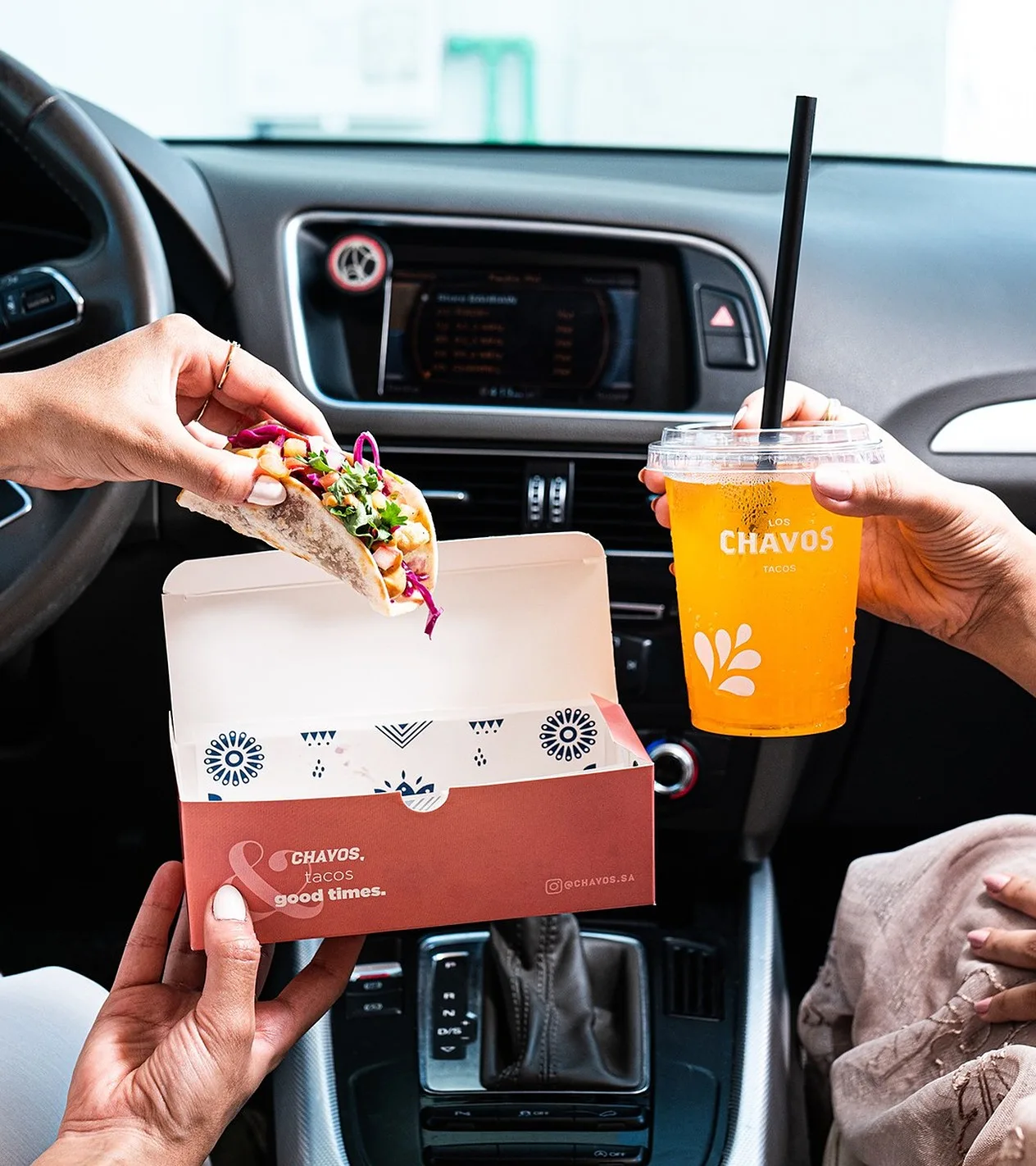 Dos manos dentro de un coche sostienen una caja de taco roja y blanca con la frase “&CHAVOS, tacos good times.” y un vaso naranja con el logotipo.