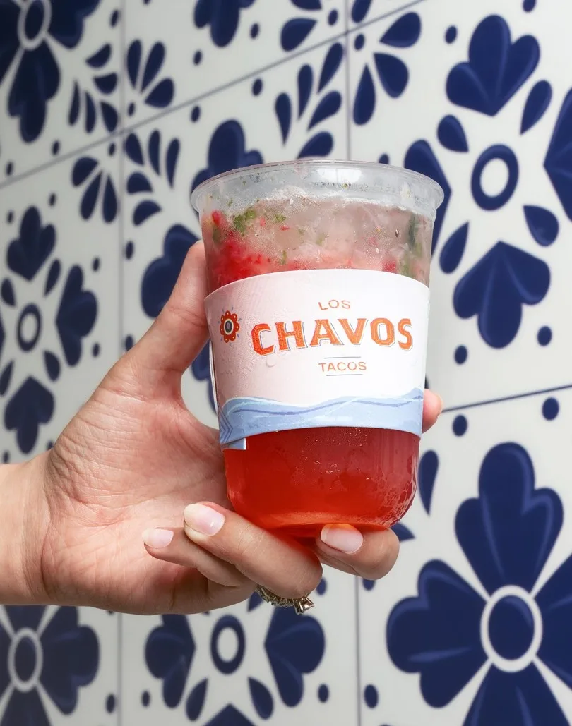 Vaso con la faja del logotipo en una mano, frente a un muro de azulejos azules y blancos.