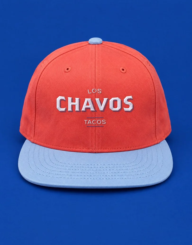 Mockup de una gorra roja con visera azul claro y el logotipo de Los Chavos bordado en blanco, sobre fondo azul rey.