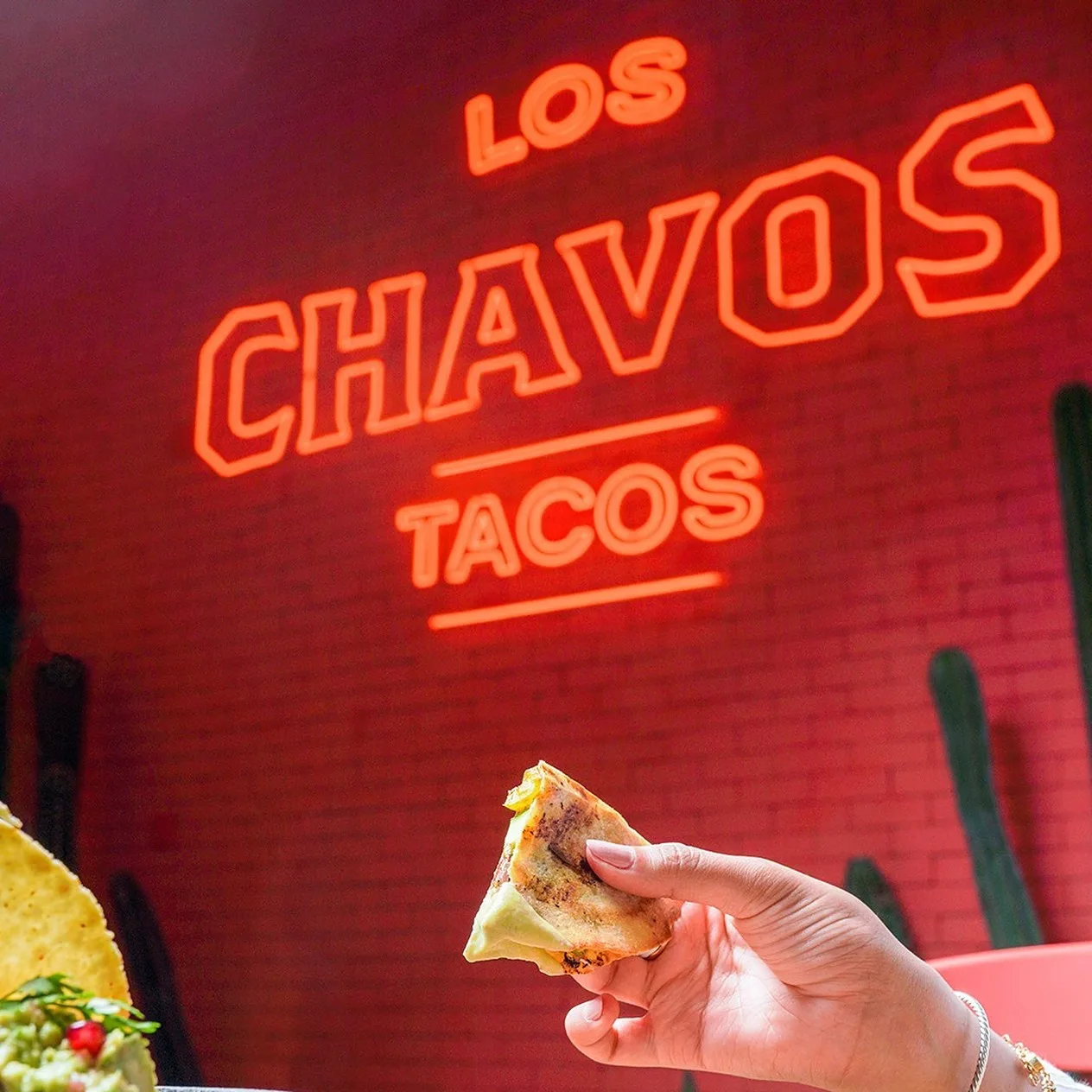 El logotipo de Los Chavos en neón rojo sobre un muro de ladrillo rojo, con una mano que sostiene una quesadilla abajo.