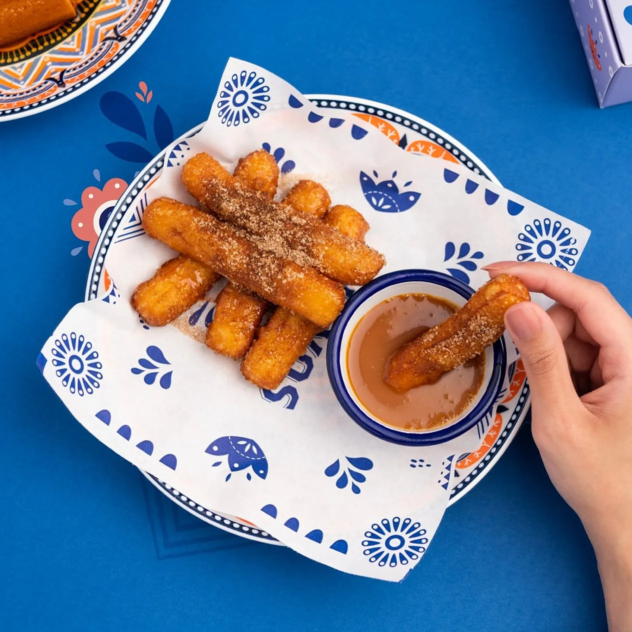Publicación de Instagram sobre azul, vista desde arriba: churros en el papel de la marca, con dibujos azules, y una mano que moja uno en la salsa.
