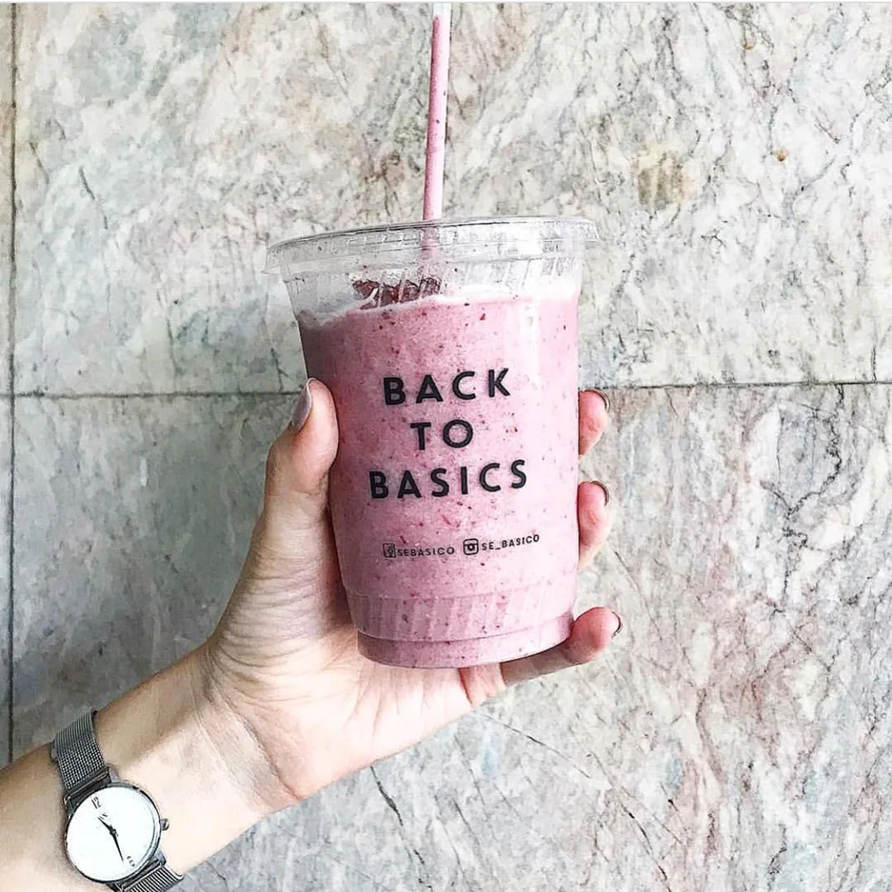 Mano que sostiene un smoothie morado en vaso transparente con «Back to Basics» impreso.