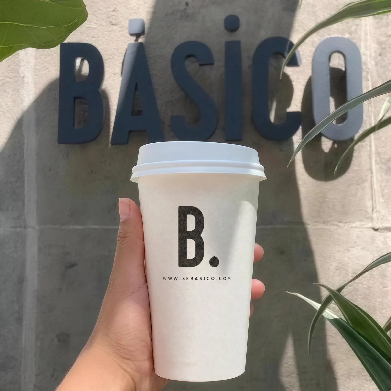 Mano que sostiene un vaso blanco de café para llevar con la B. negra y sebasico.com, delante del letrero BÁSICO.